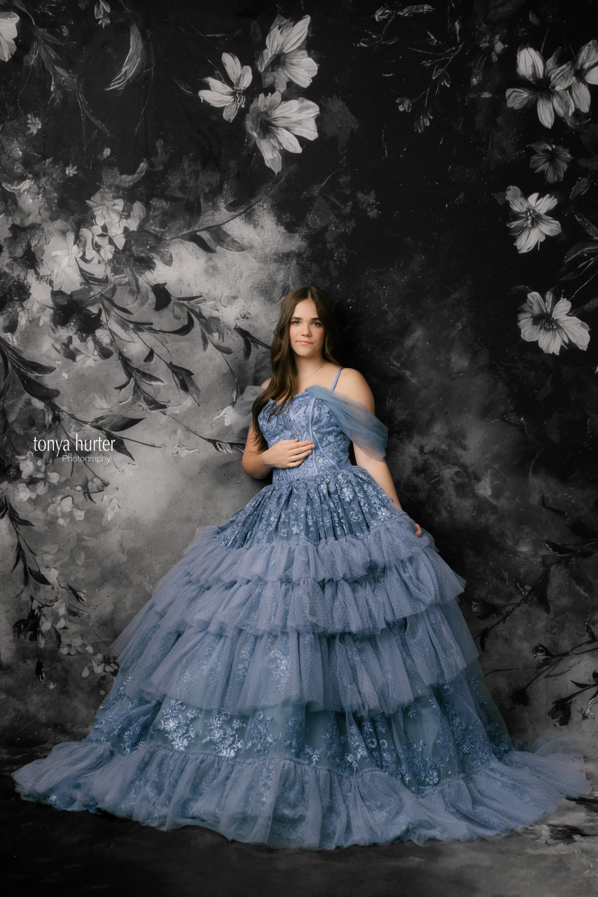 Woman in a blue tiered dress standing against a dark floral backdrop