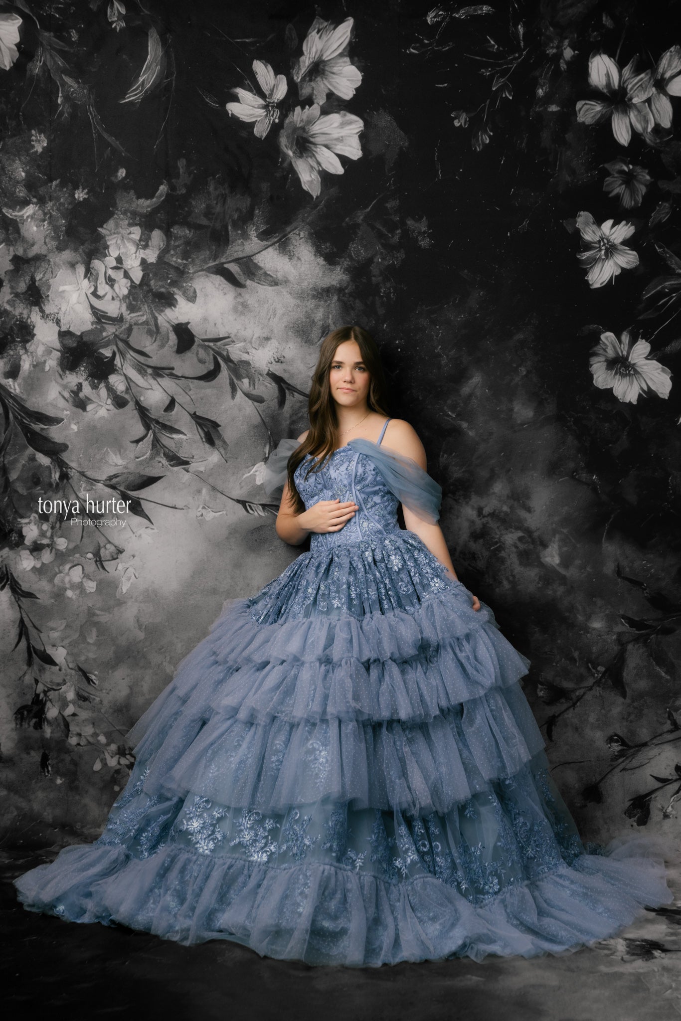 Woman in a blue tiered dress standing against a dark floral backdrop