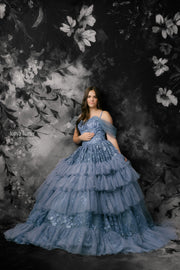 Woman in a blue tiered dress standing against a dark floral backdrop