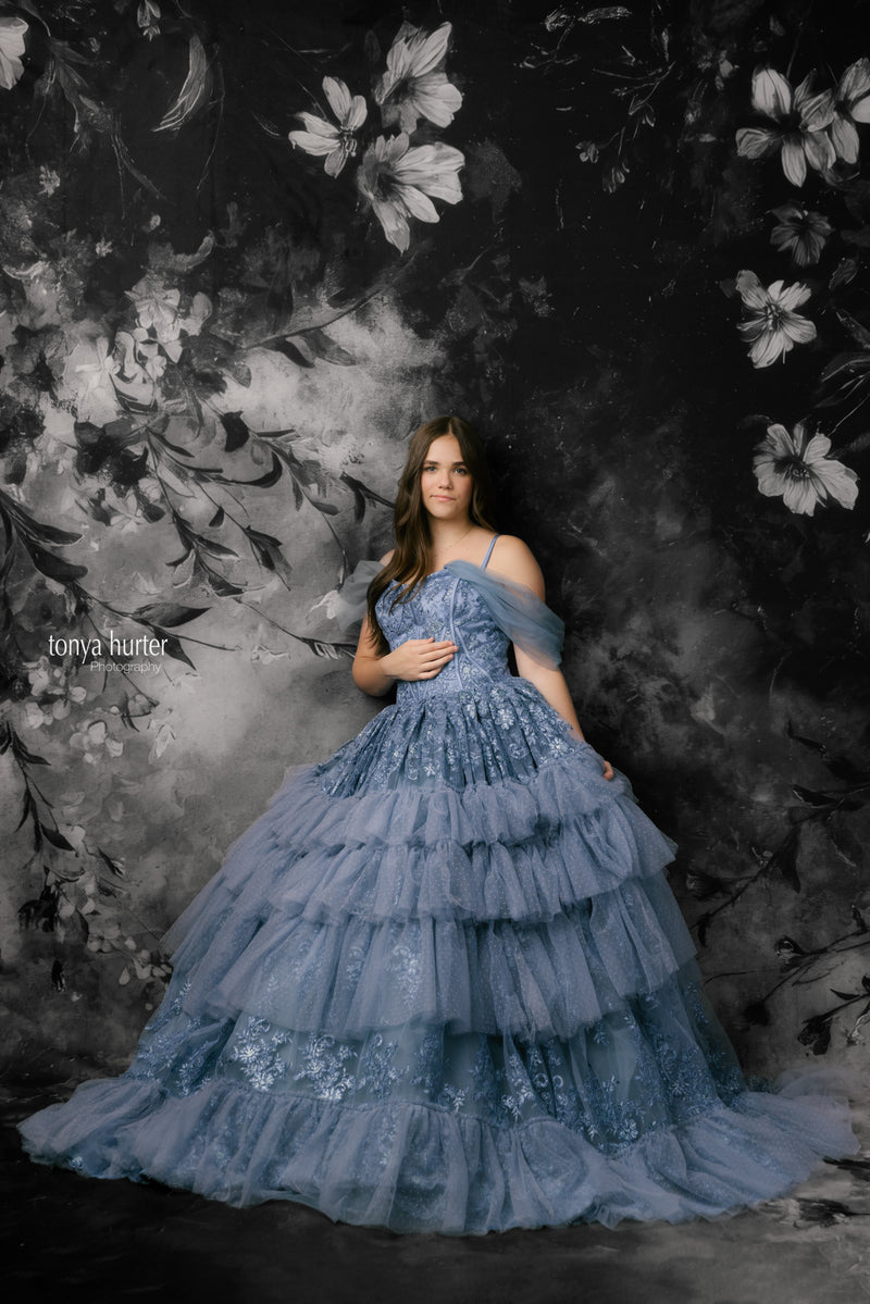 Woman in a blue tiered dress standing against a dark floral backdrop