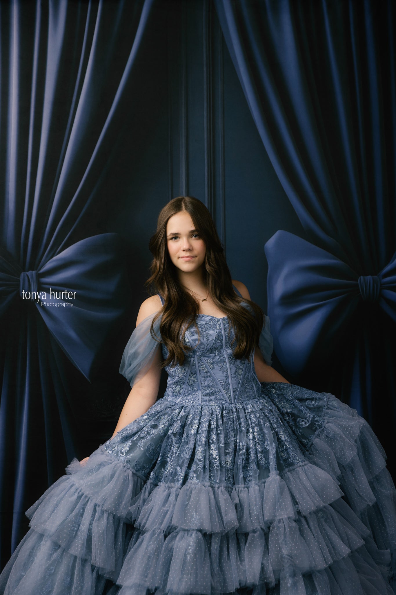 Woman in a blue dress sitting against a dark curtain with large blue bows