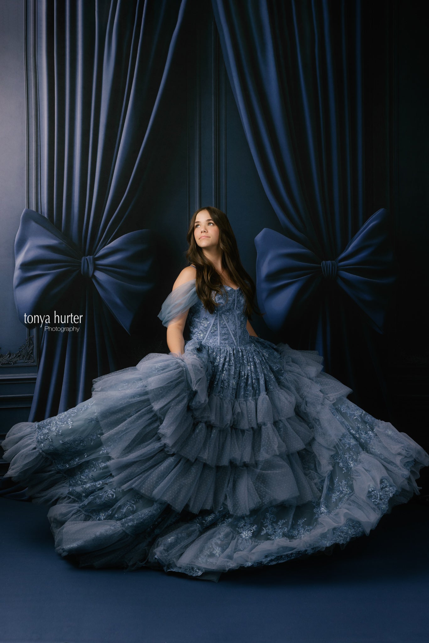 Woman in a blue gown standing in front of dark blue curtains with large bows.