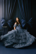 Woman in a blue gown standing in front of dark blue curtains with large bows.