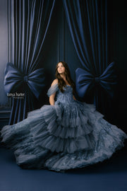 Woman in a blue gown standing in front of dark blue curtains with large bows.