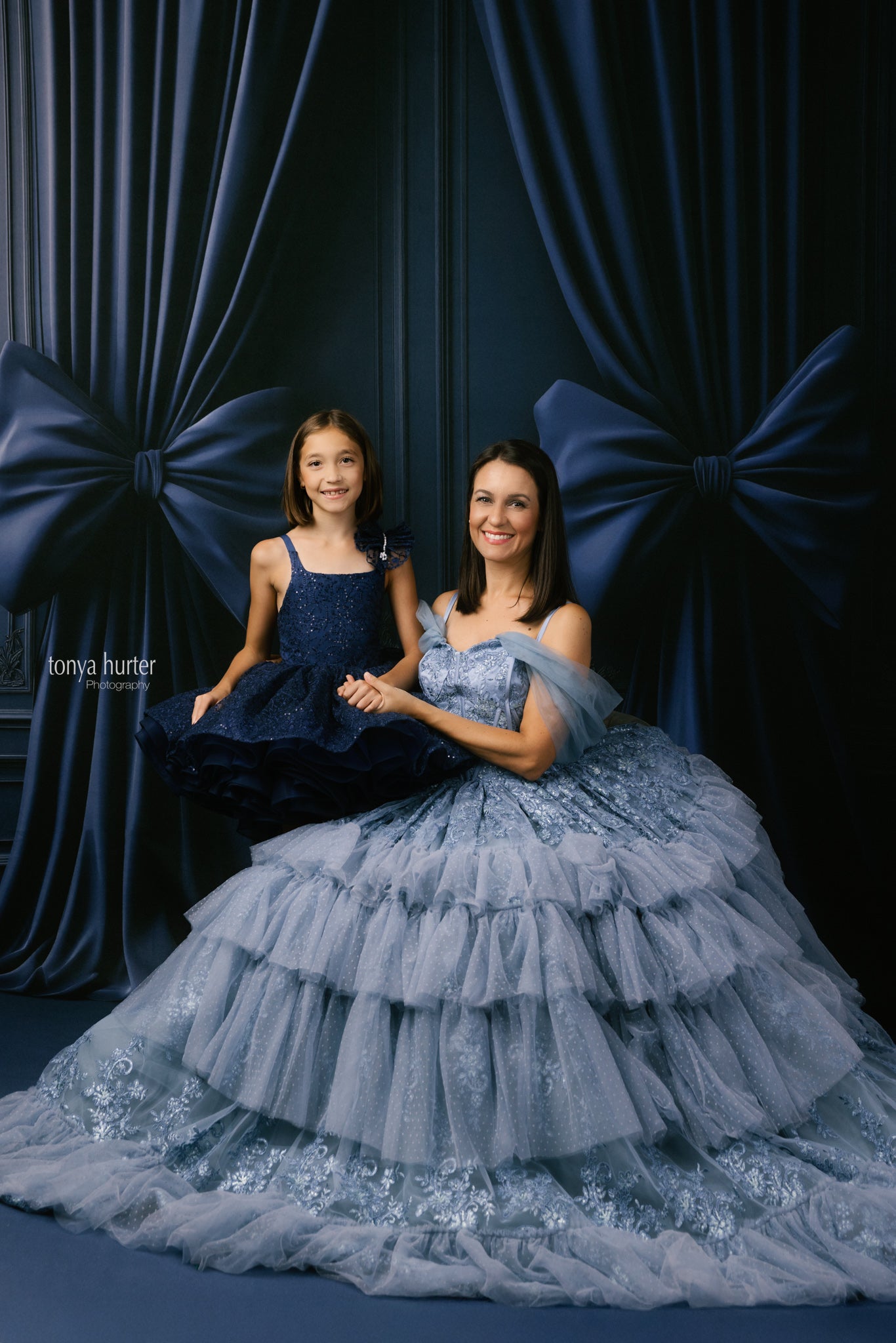 Two women in elegant dresses sitting against a dark curtain backdrop