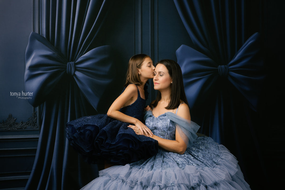 Two women in formal dresses sitting close together against a dark blue curtain with large bows.