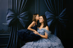 Two women in formal dresses sitting close together against a dark blue curtain with large bows.