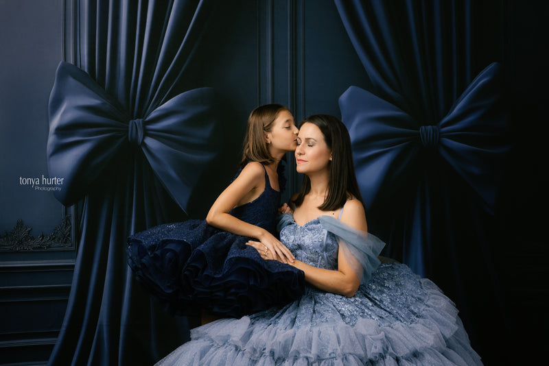 Two women in formal dresses sitting close together against a dark blue curtain with large bows.