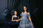 Two women in formal dresses standing in front of dark blue curtains with a bow.