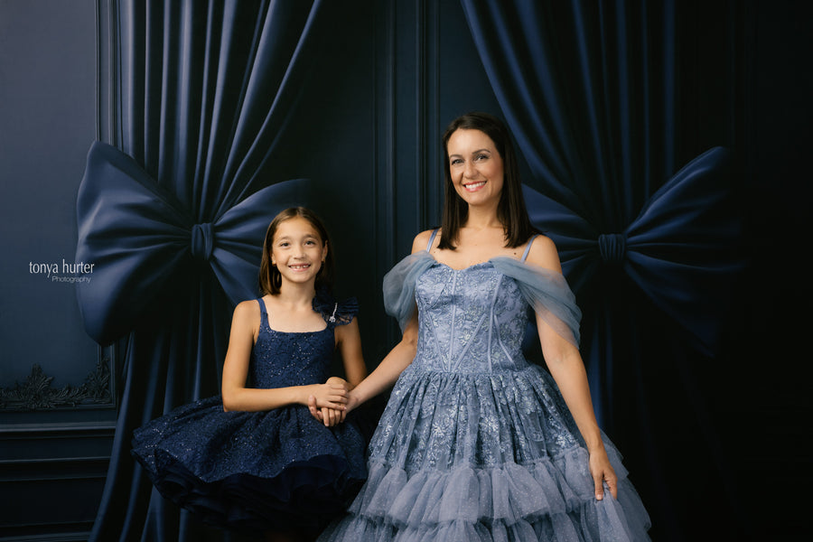 Two women in formal dresses standing in front of dark blue curtains with a bow.