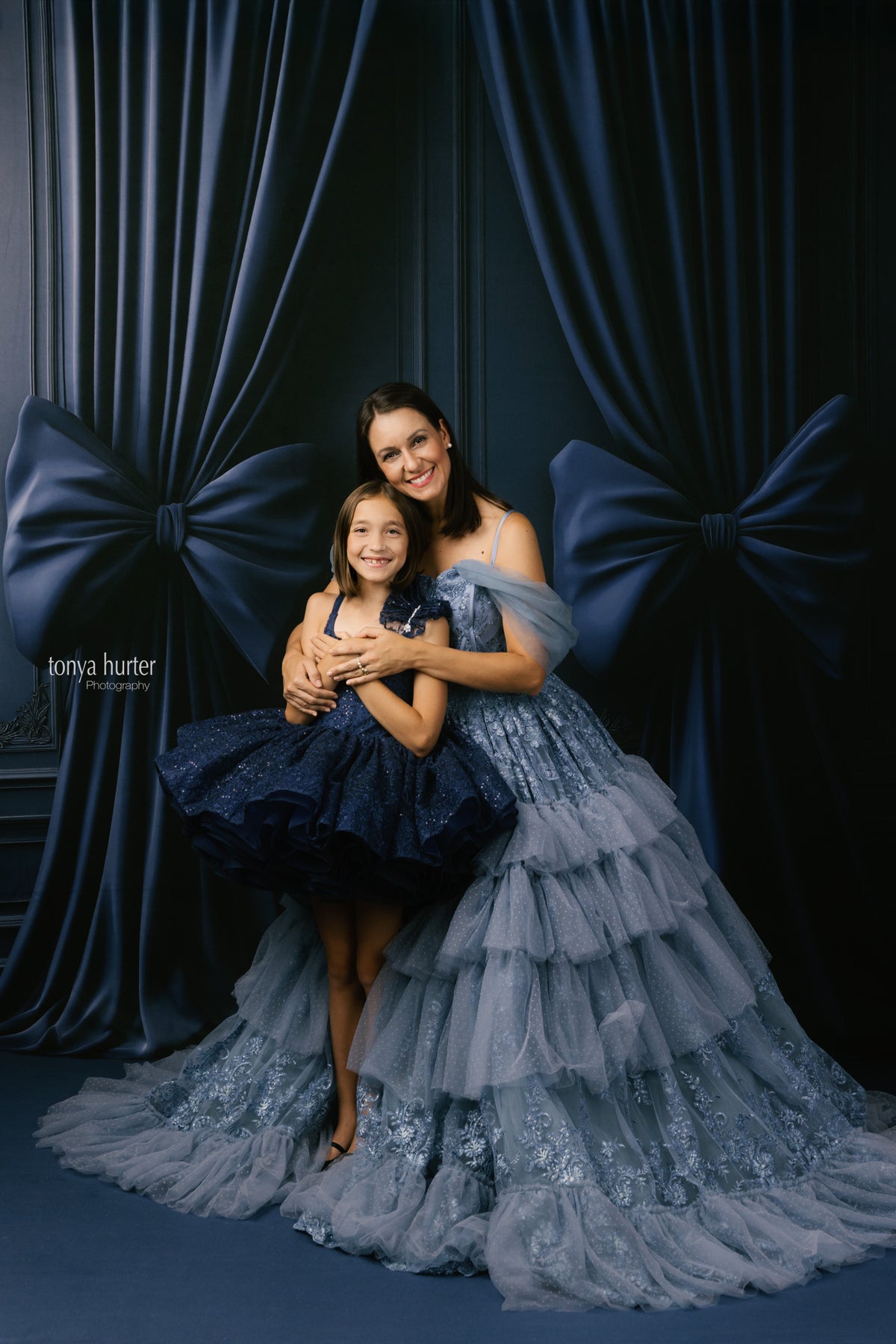 Two women in elegant dresses posing against a dark curtain backdrop
