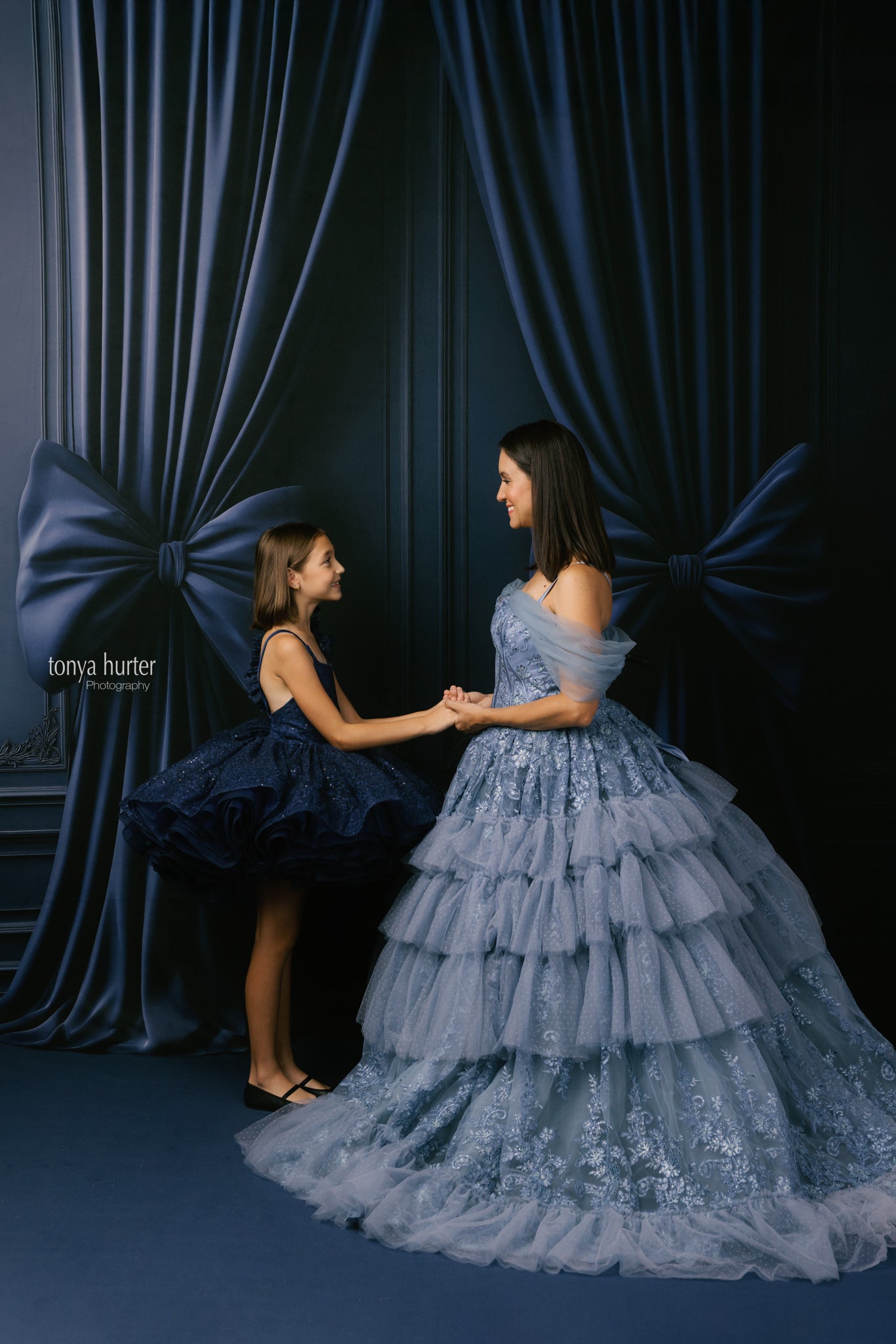 Two women in formal dresses standing in front of a dark curtain with large blue bows.