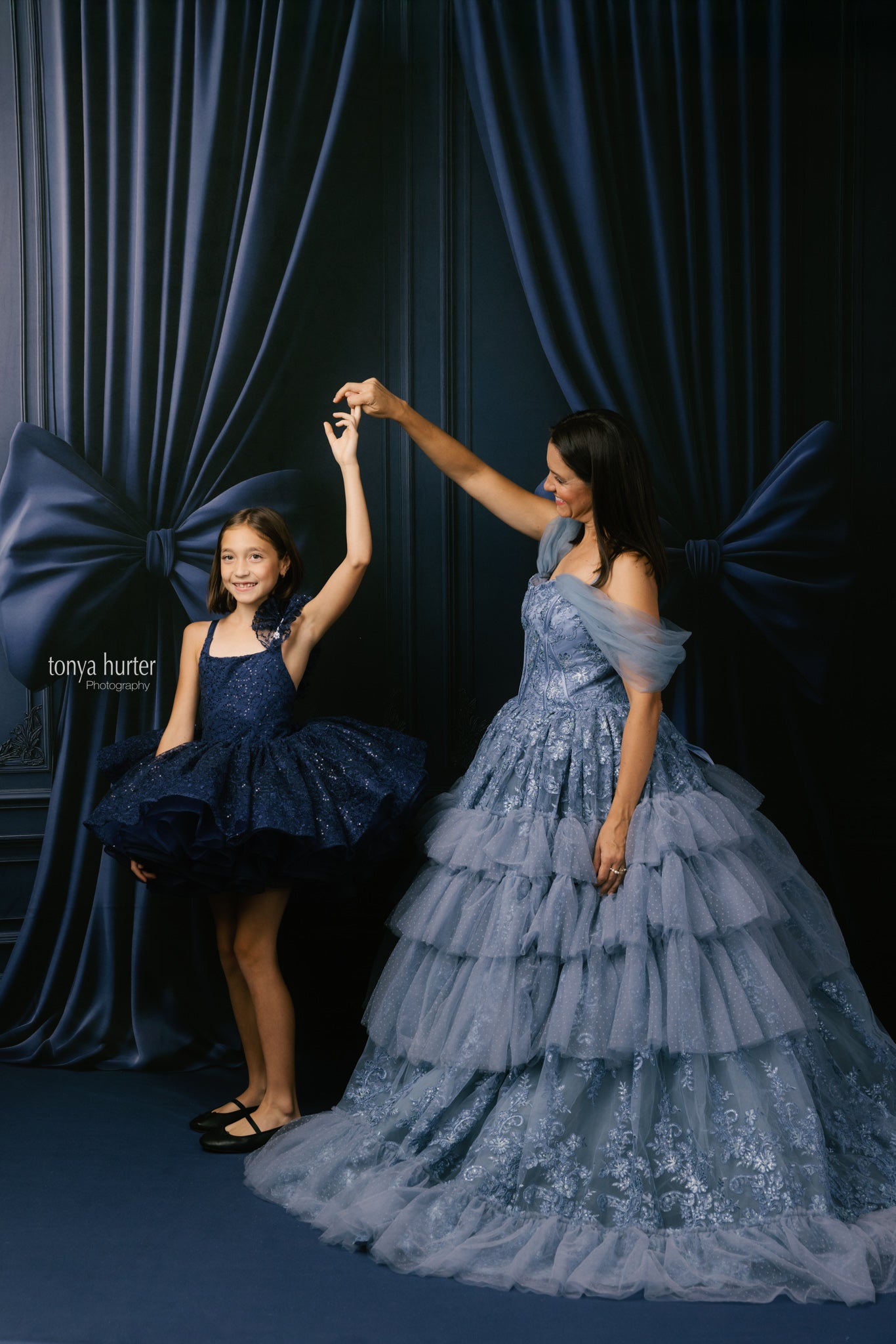 Woman and young girl in formal dresses posing against a dark curtain backdrop