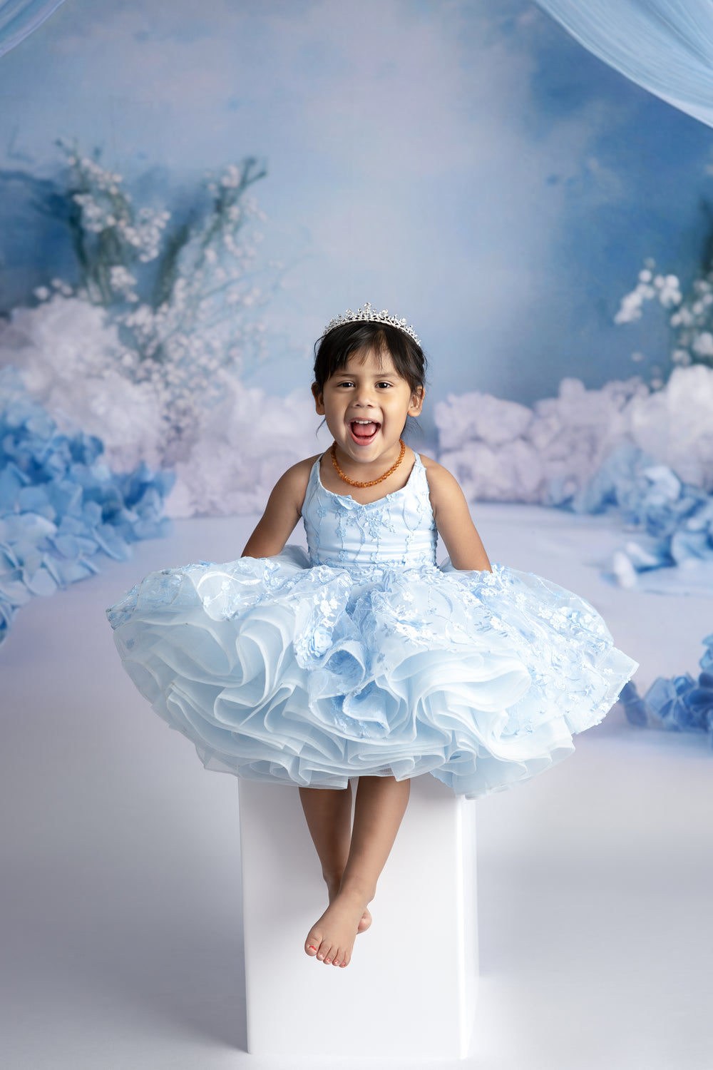 Young girl wearing Bentley + Lace’s Heavenly Petal gown in soft blue, standing in soft natural light, highlighting the 3D floral details and flowing tulle.

