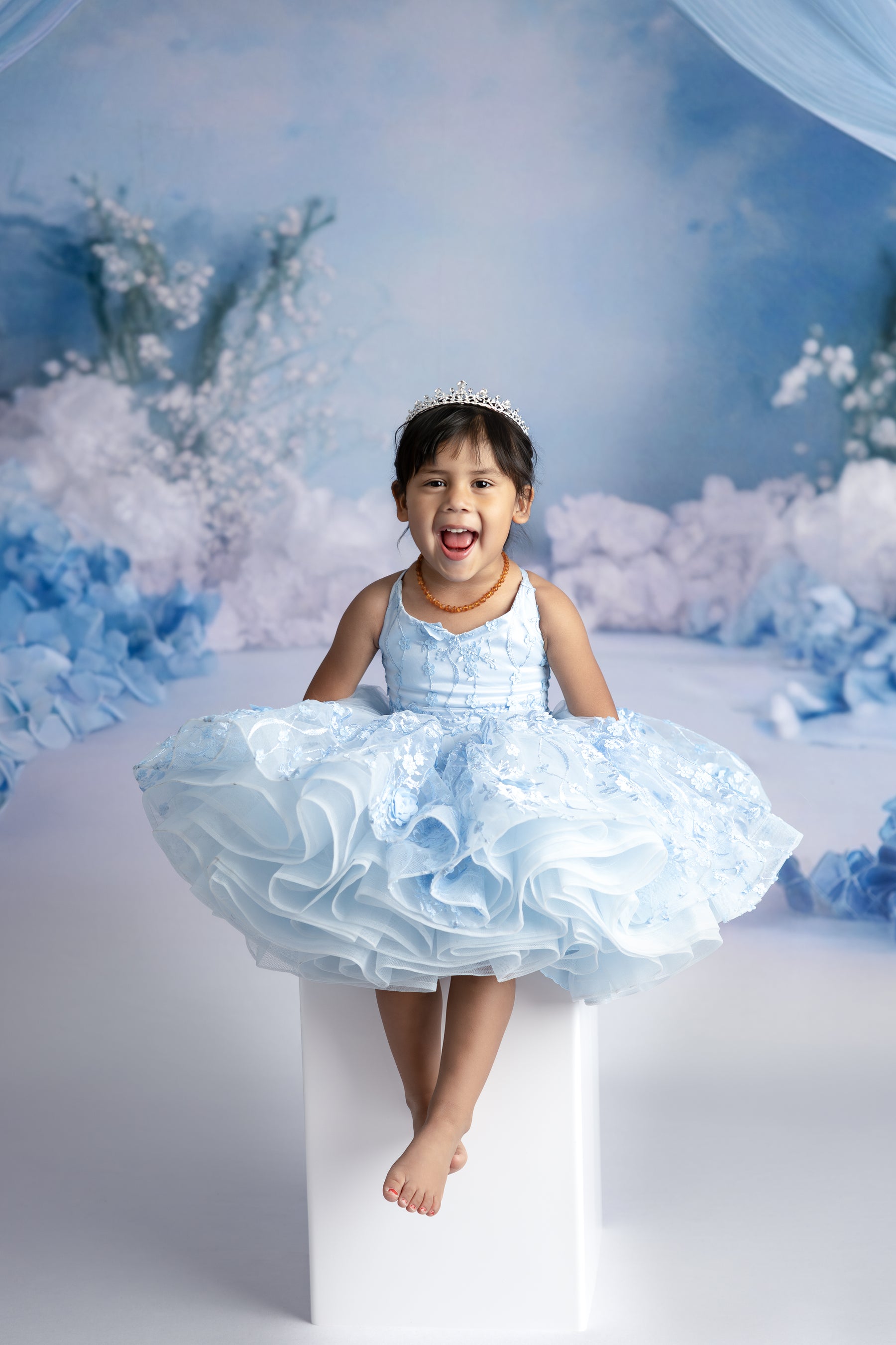 Young girl wearing Bentley + Lace’s Heavenly Petal gown in soft blue, standing in soft natural light, highlighting the 3D floral details and flowing tulle.

