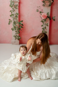 Infant dressed in The Rosalie Romper, showcasing soft cream velvet, intricate embroidered lace, and gathered silhouette for comfort and elegance.

