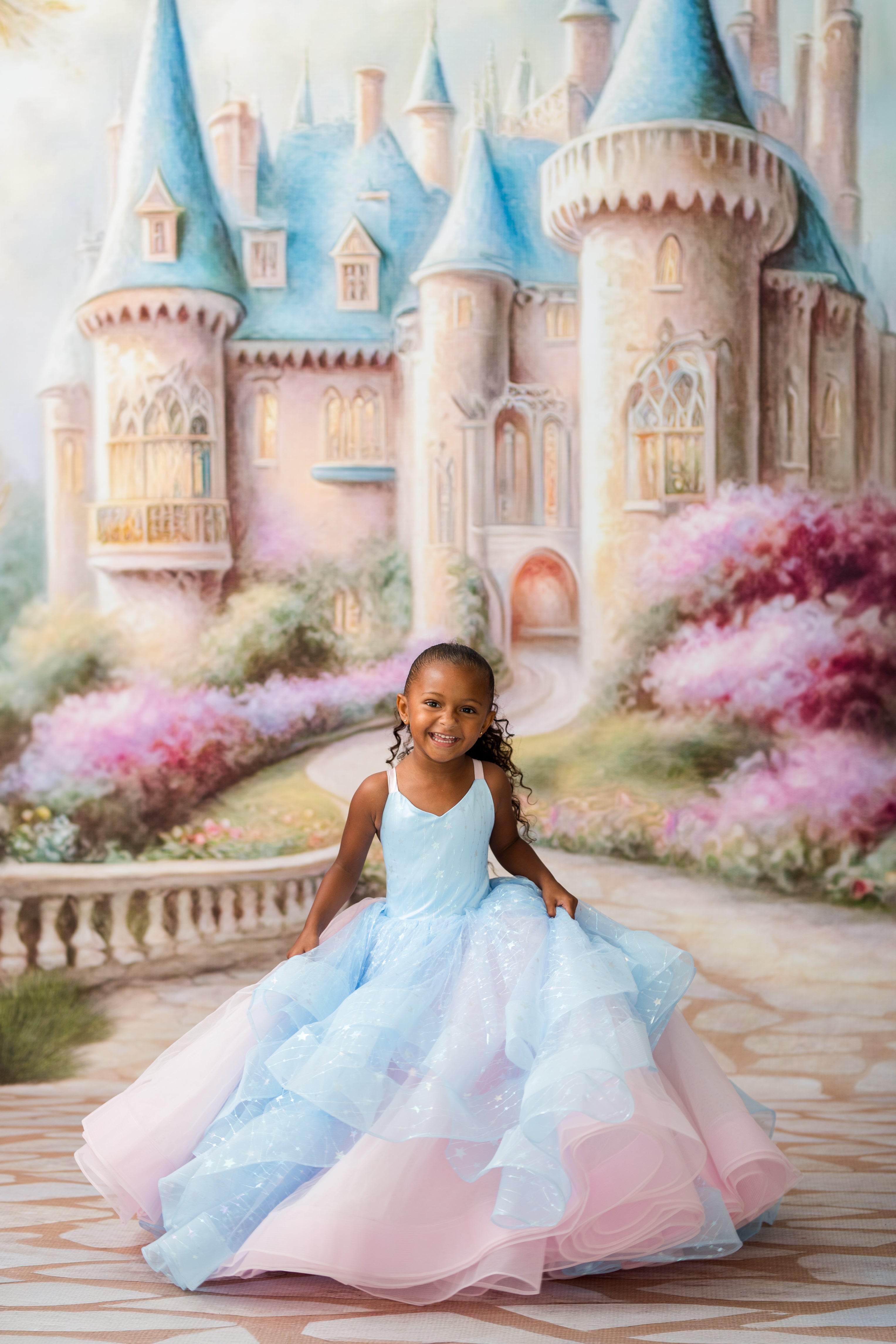 A young girl with braided hair sits on a stone path in front of a painted fairytale castle, wearing a voluminous ball gown with a light blue bodice and layered tulle skirt in pale blue and pink.
