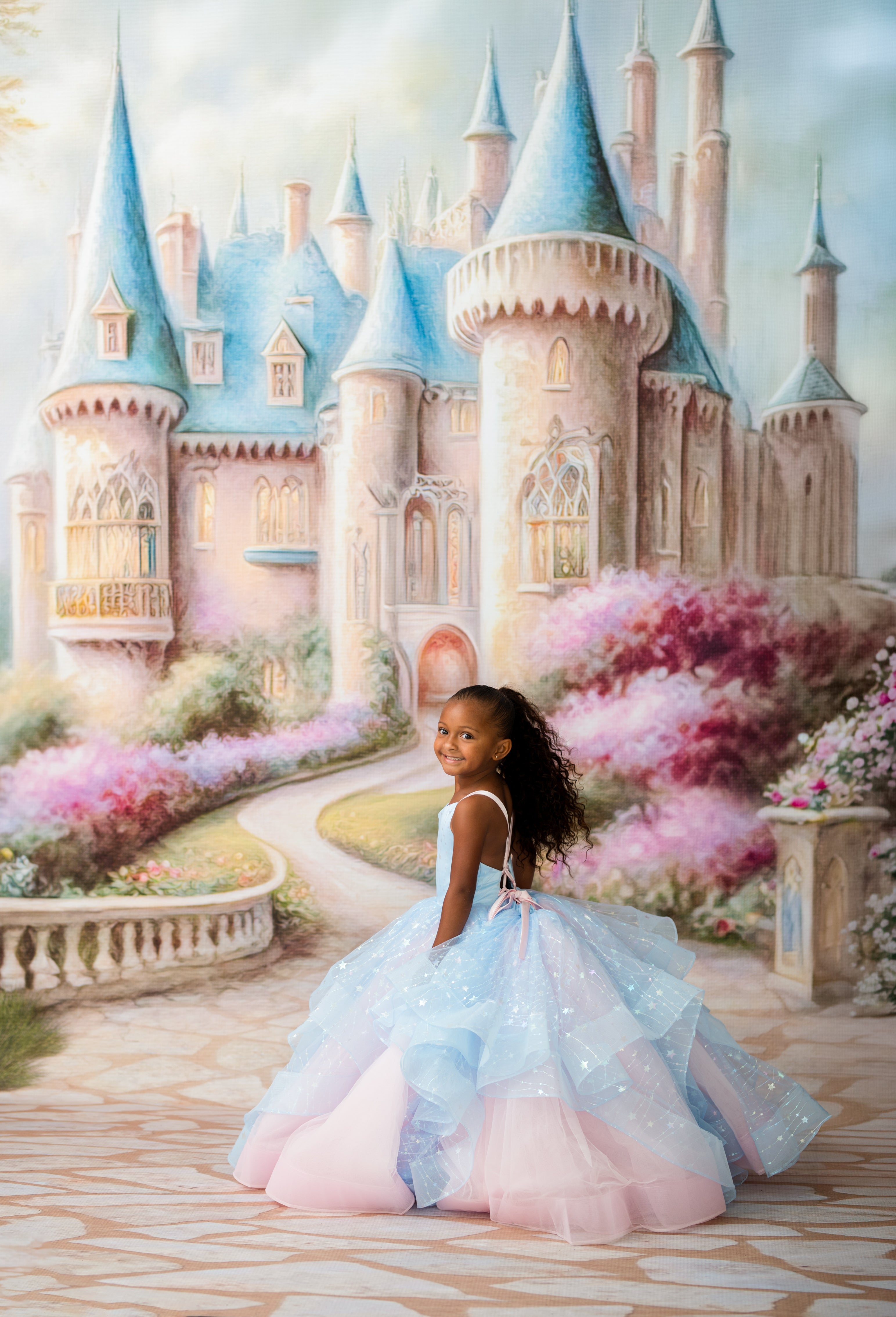 A young girl in a pastel blue and pink ballgown stands in front of a whimsical painted castle with blue spires and pink flowering bushes.