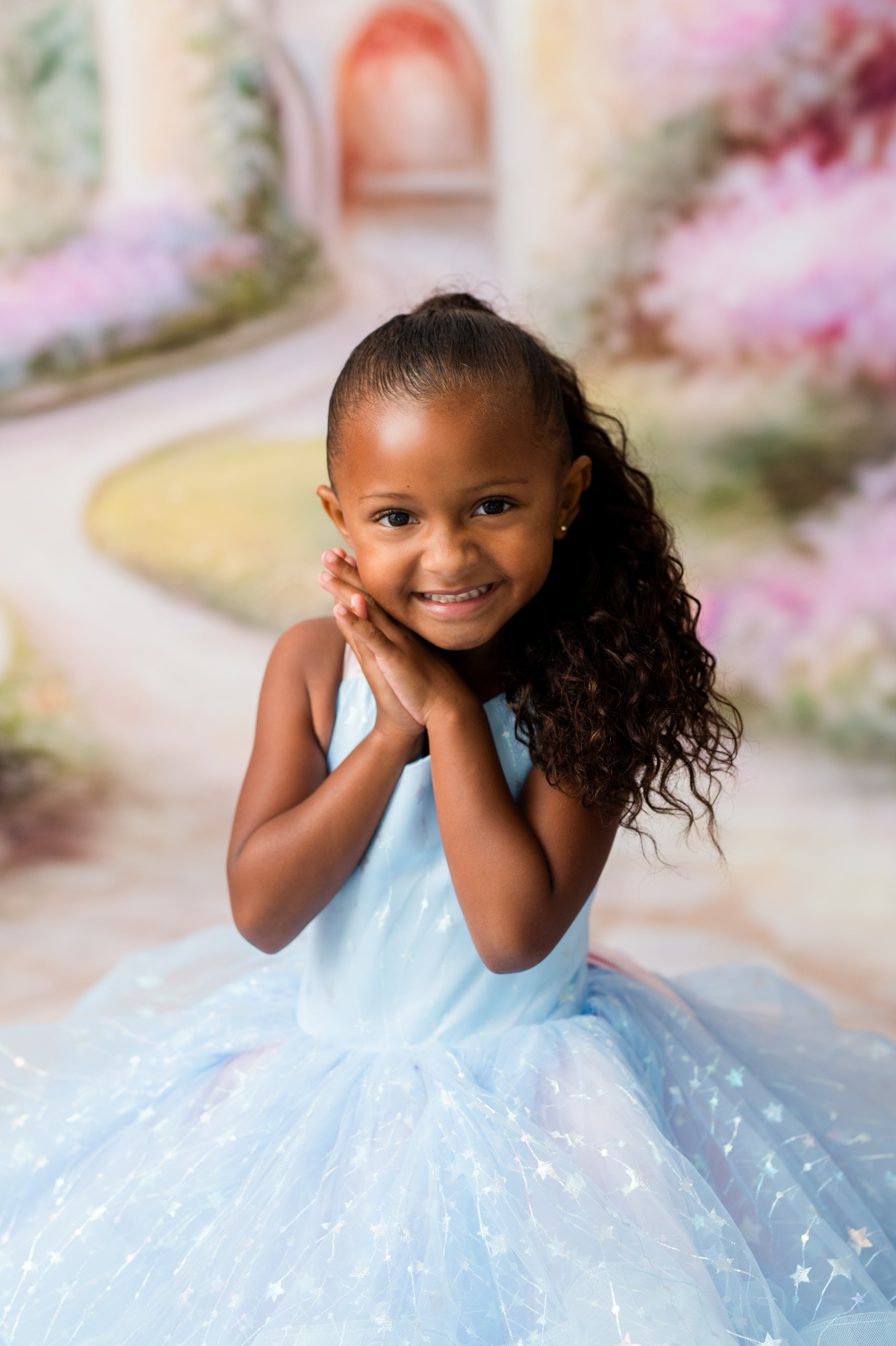 A young girl with curly brown hair wearing a light blue sparkly tulle ball gown, posing with her hands together in front of a soft, blurred fantasy garden backdrop.