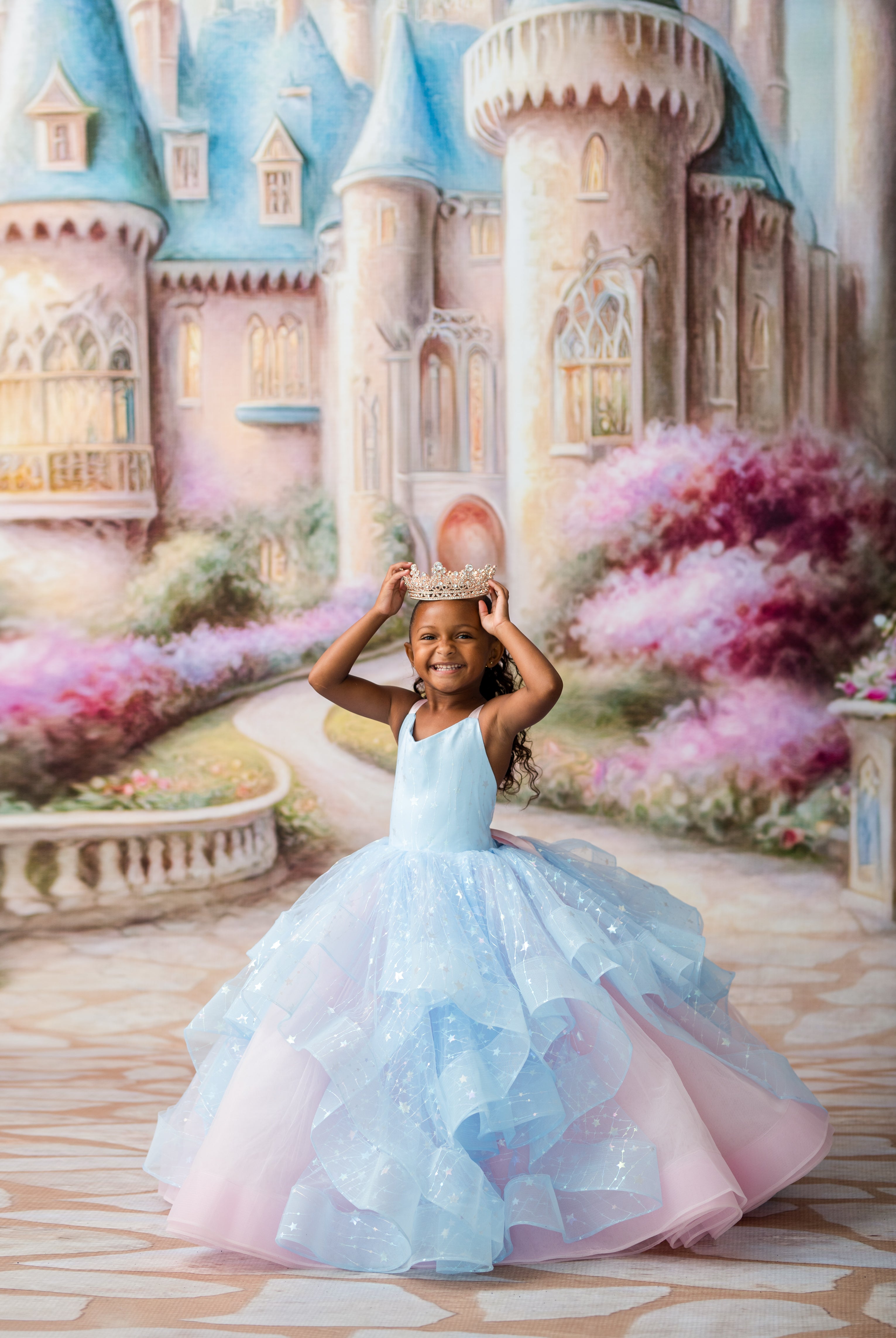 A young girl wearing a light blue and pink glittery ball gown with a tiara, posing in front of a painted castle backdrop.