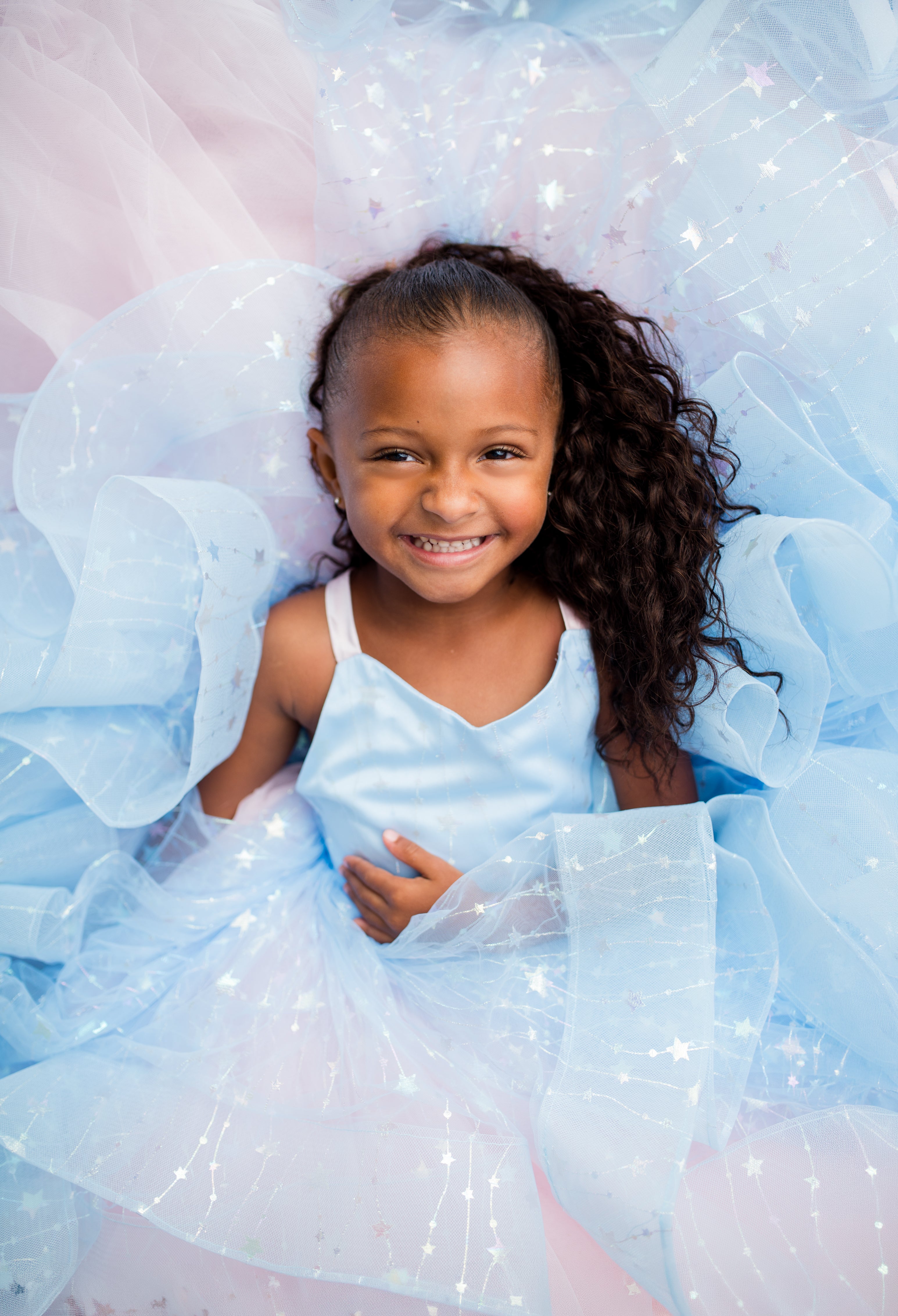 A young girl with long curly brown hair smiling while wearing a light blue satin top and a voluminous light blue tulle skirt with sparkly details, posed on a soft pink and blue tulle background.