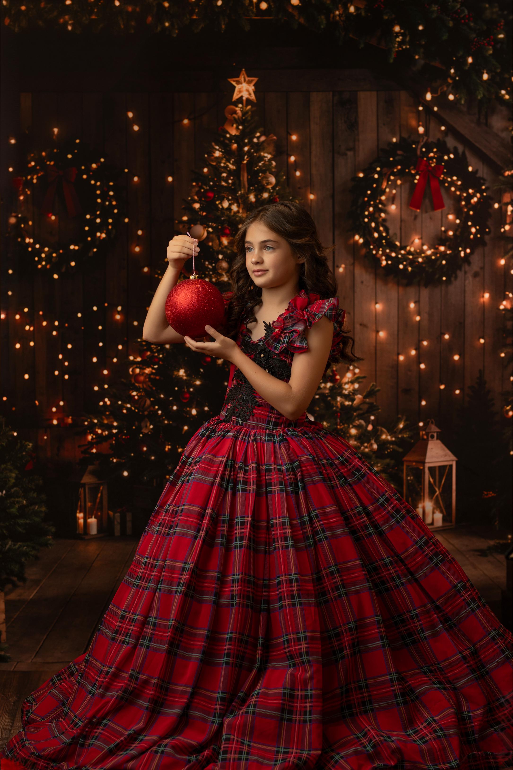 Smiling young girl in a red tartan holiday gown, posing in front of a Christmas tree glowing with warm lights.
