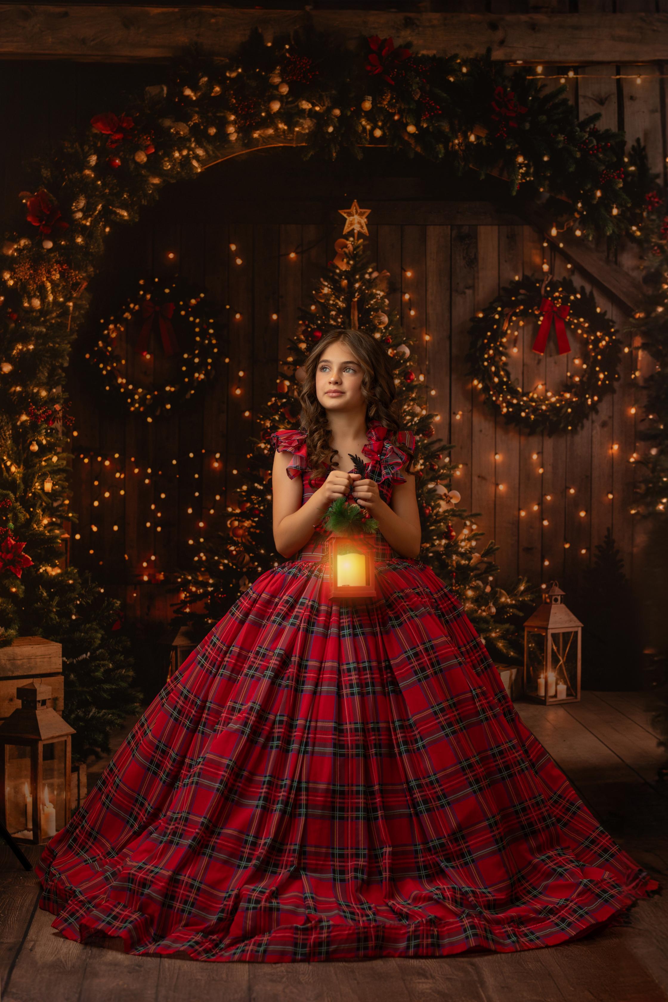 Child wearing a festive plaid holiday dress, holding out the skirt, with a Christmas tree decorated with lights in the background.
