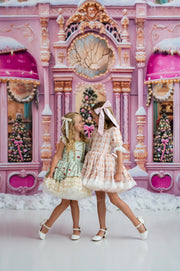 Two young girls in matching pink holiday-themed dresses pose together in front of a whimsical pink Christmas village backdrop with snow-covered ground.