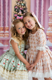 Two young girls wearing matching vintage-style dresses with floral patterns, standing together in front of a pink holiday backdrop with a decorated Christmas tree.