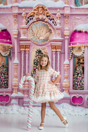 A young girl stands in front of a whimsical pink fantasy backdrop resembling a snowy castle or fairground, holding a large striped candy cane. She is wearing a white and pink patterned dress with white shoes.