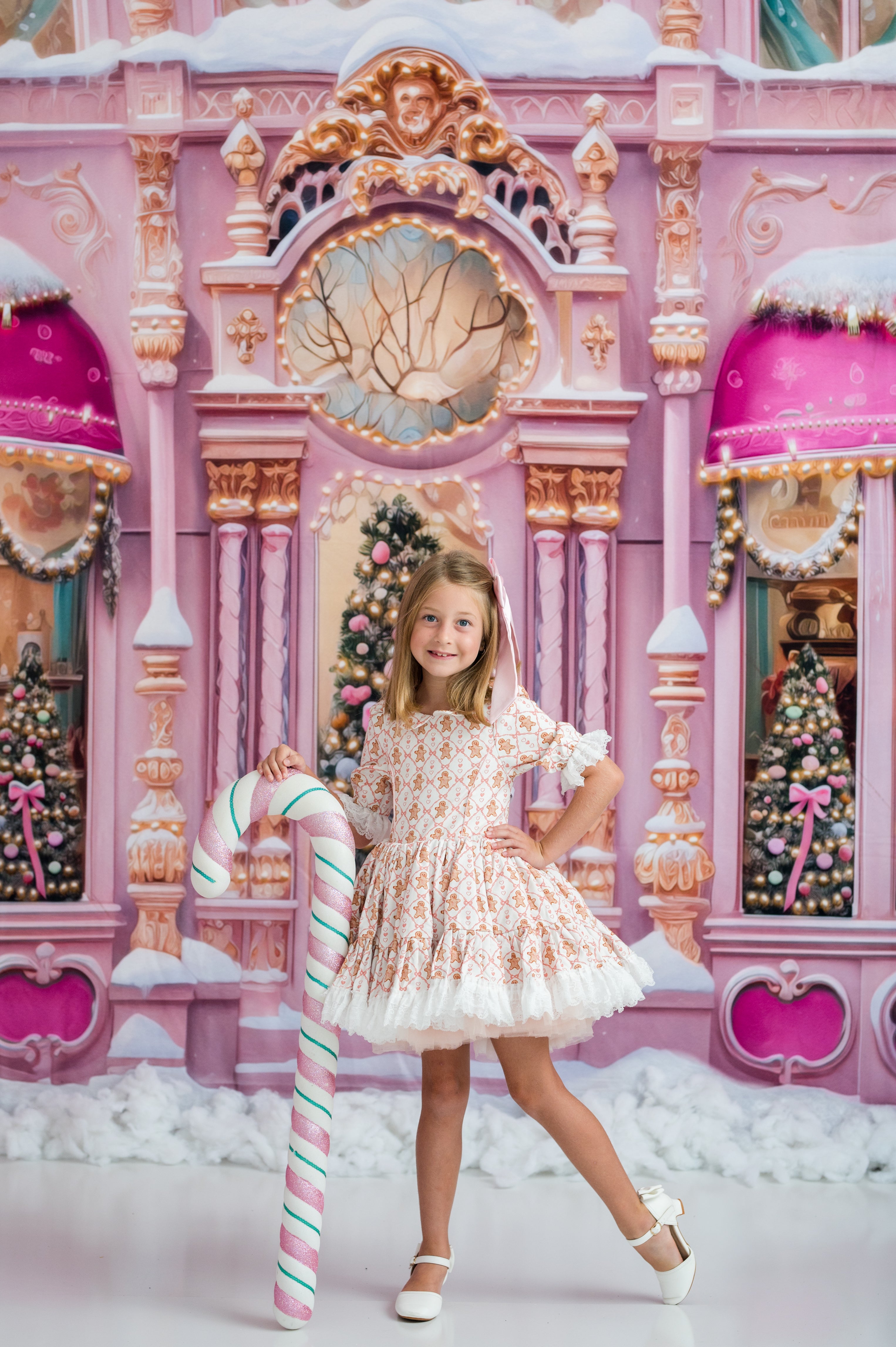 A young girl stands in front of a whimsical pink fantasy backdrop resembling a snowy castle or fairground, holding a large striped candy cane. She is wearing a white and pink patterned dress with white shoes.