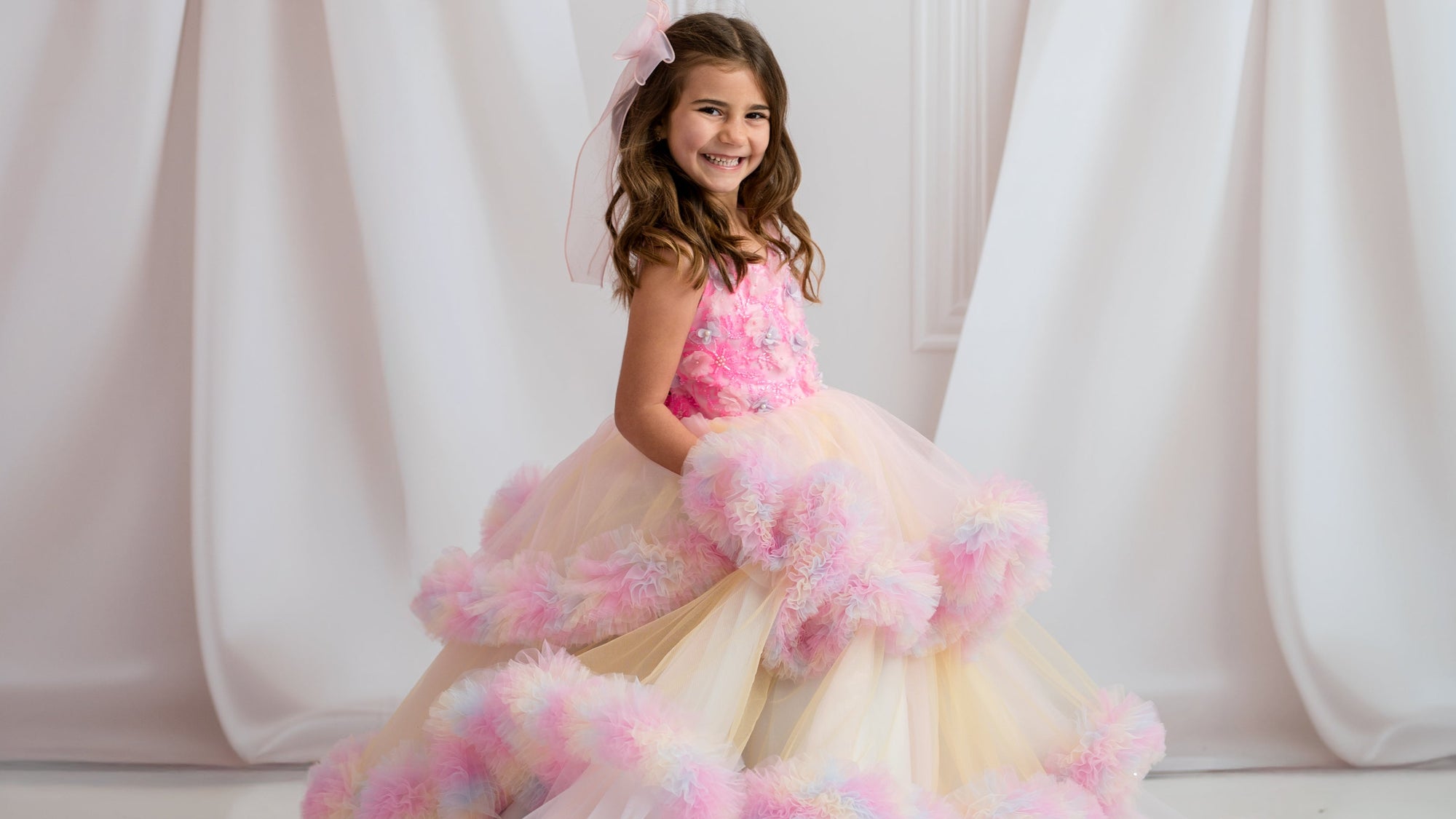 Young girl in a pink and white dress with floral details against a decorative white curtain background.