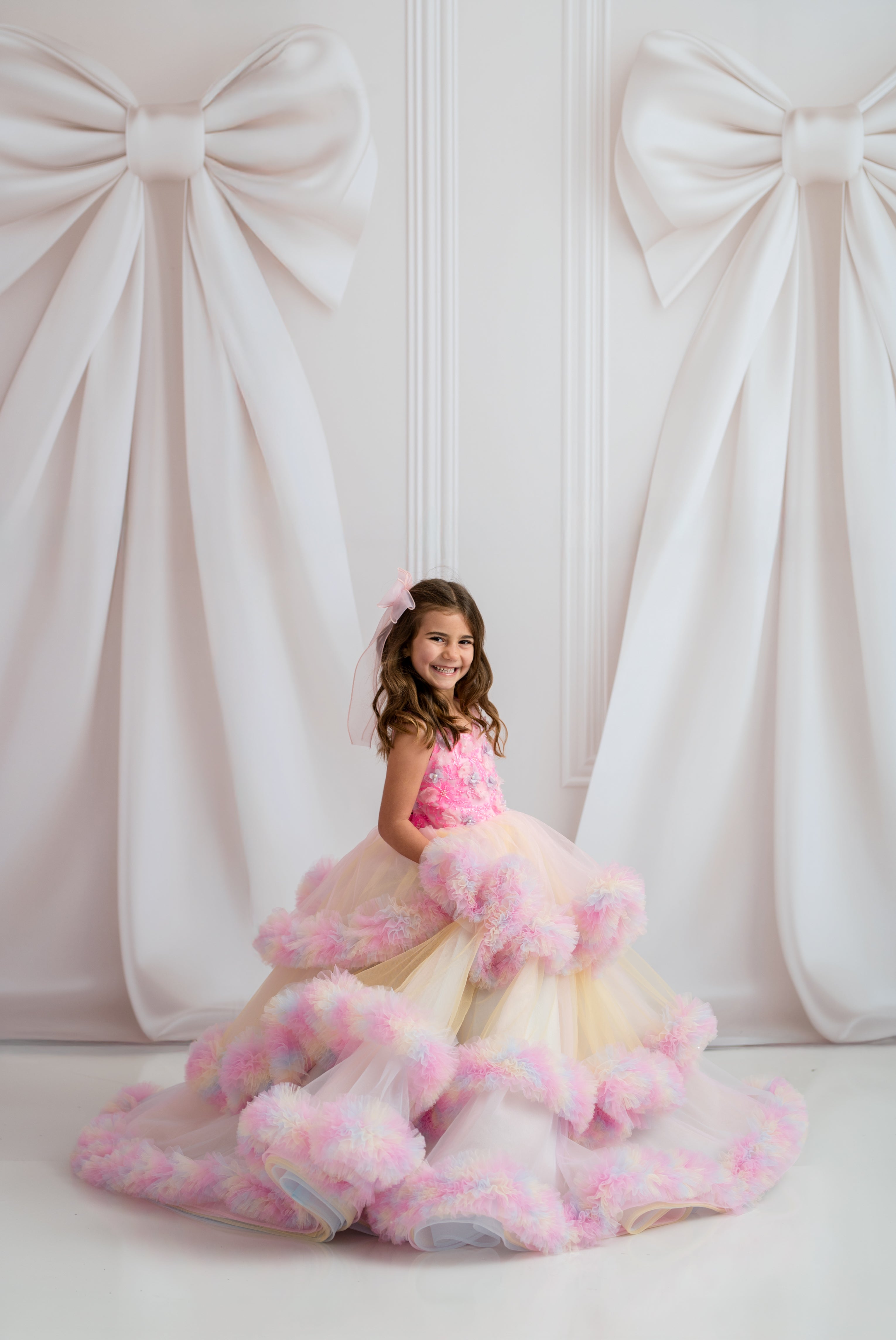 Young girl in a pink and white dress with floral details against a decorative white curtain background.