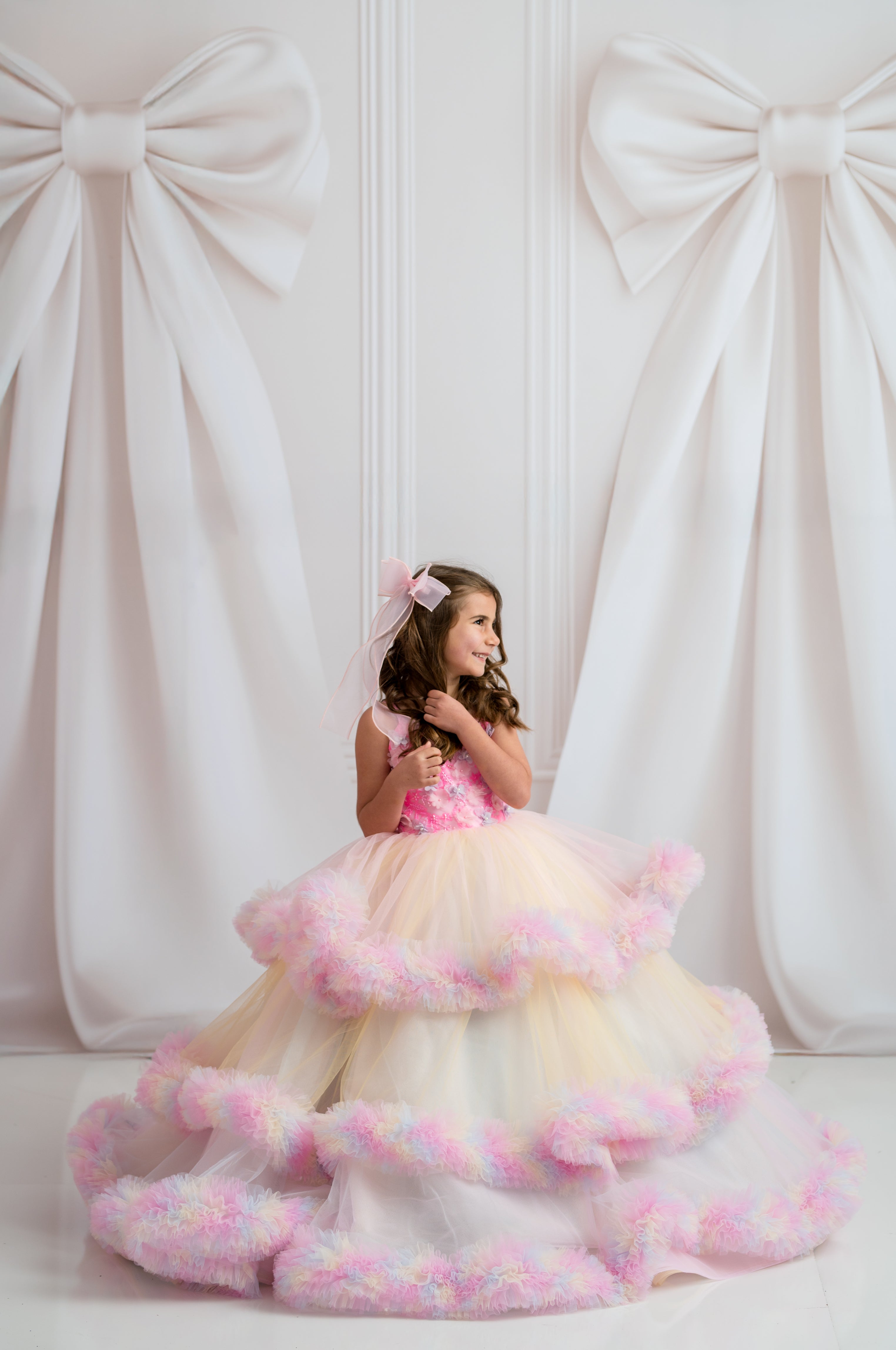 Young girl in a pink and white dress with a bow in her hair, standing against a white curtain backdrop.