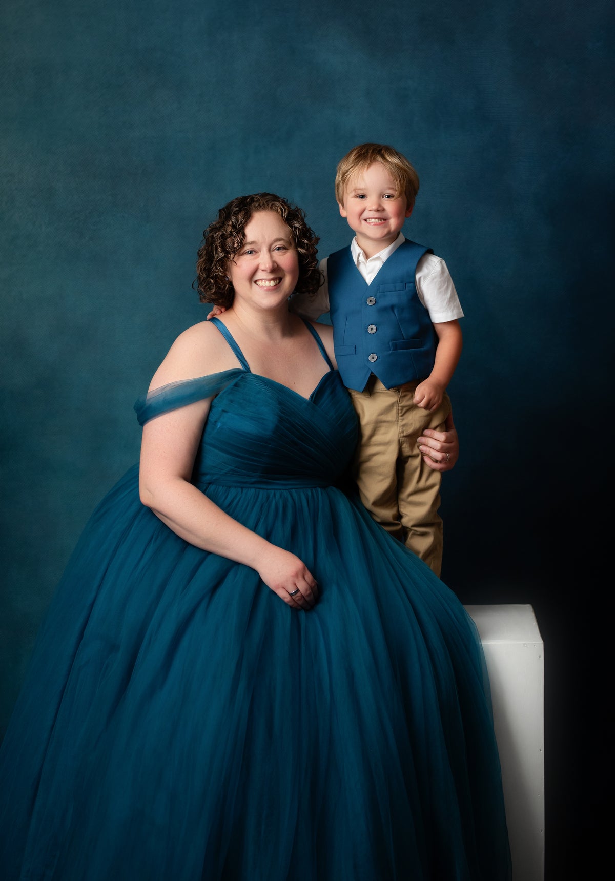 A woman in a voluminous teal off-the-shoulder gown stands beside a young boy wearing a matching teal vest over a white shirt and tan pants, posed against a dark blue studio backdrop.