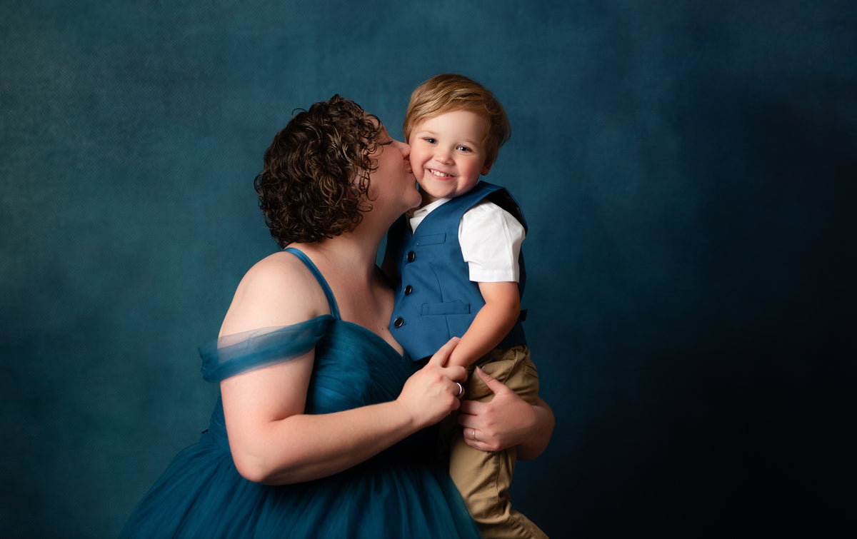 A woman in a teal blue off-the-shoulder tulle dress kisses a young boy on the cheek as he is held. The boy is wearing a blue vest over a white shirt and tan pants, set against a dark blue textured studio backdrop.