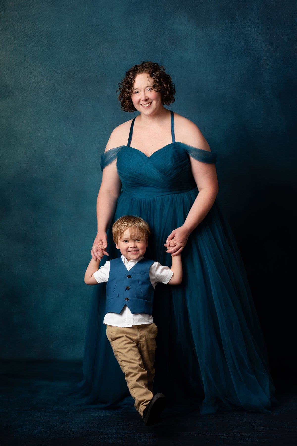 A woman in a floor-length teal off-the-shoulder gown stands behind a young boy wearing a matching teal vest and tan pants, holding his hands against a dark blue studio backdrop.