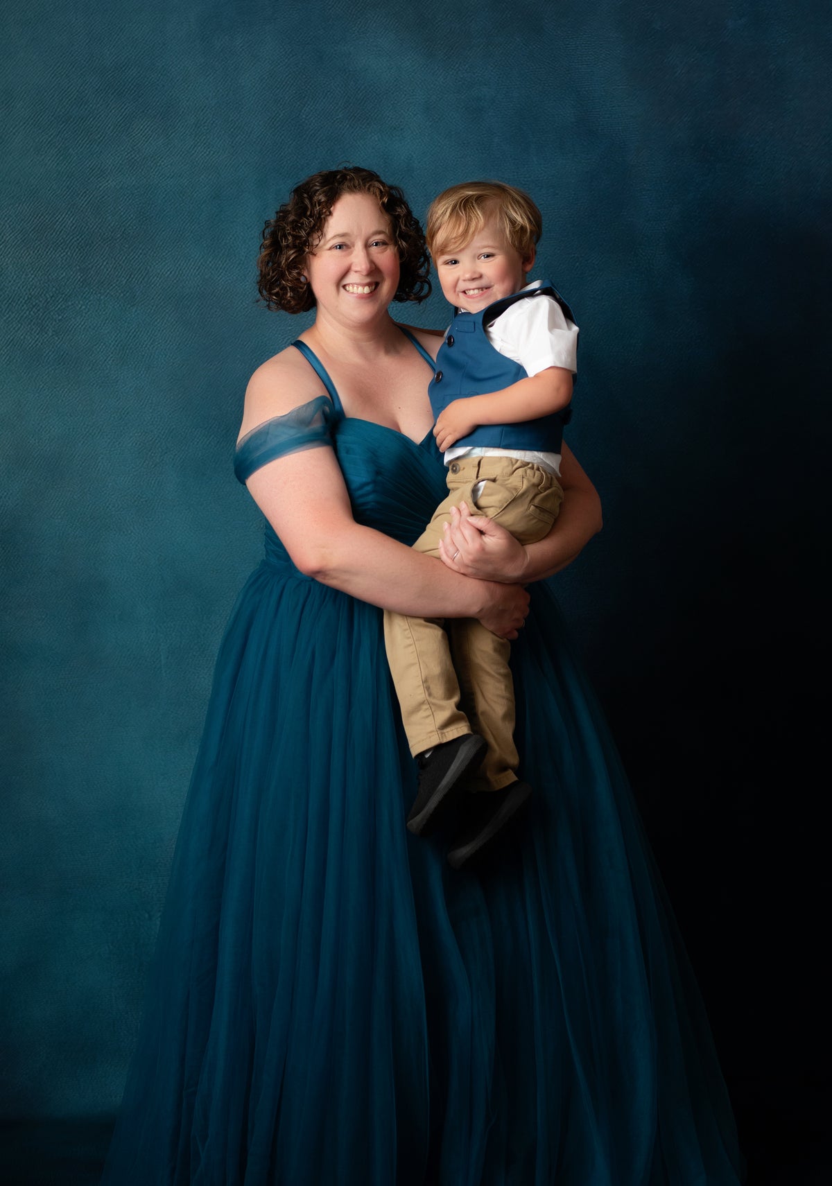 A woman in a floor-length teal blue gown holds a young boy wearing a navy vest, white shirt, and tan pants against a dark teal studio backdrop.