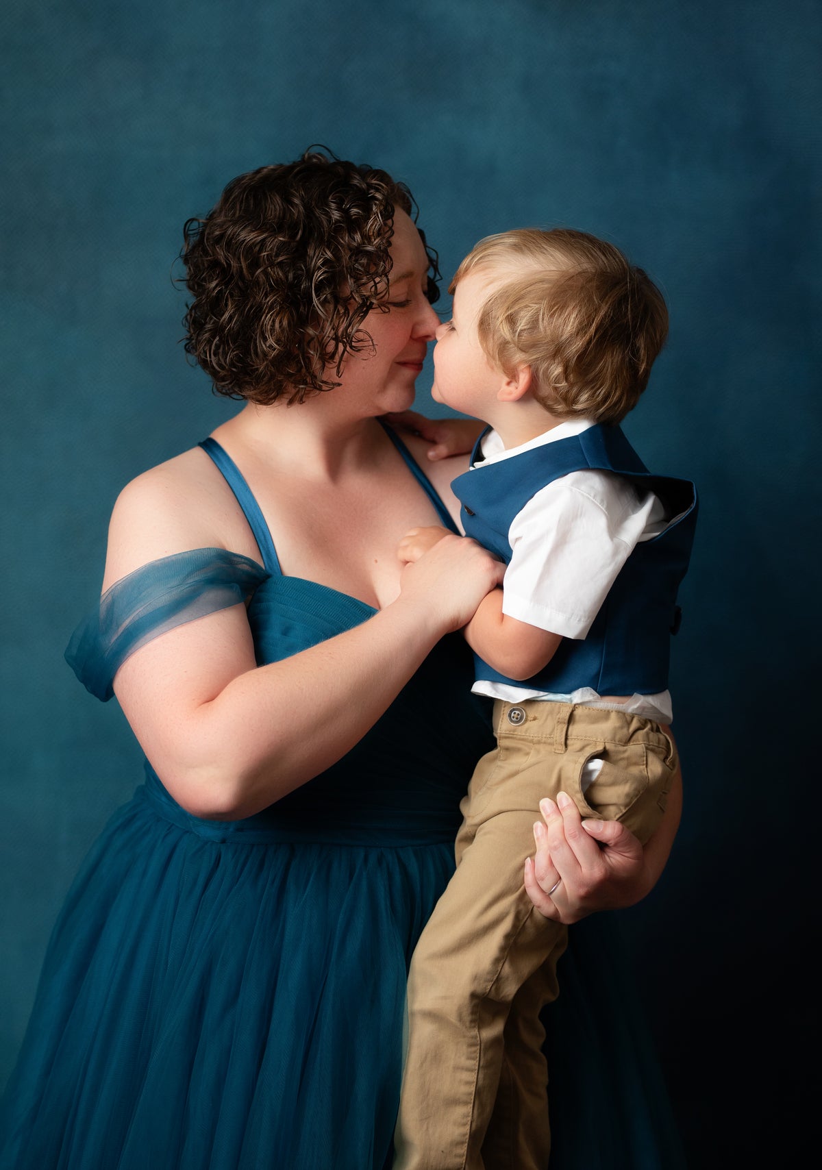 A woman in a teal blue off-the-shoulder dress holds a young child dressed in a navy vest, white shirt, and tan pants against a dark blue studio backdrop.