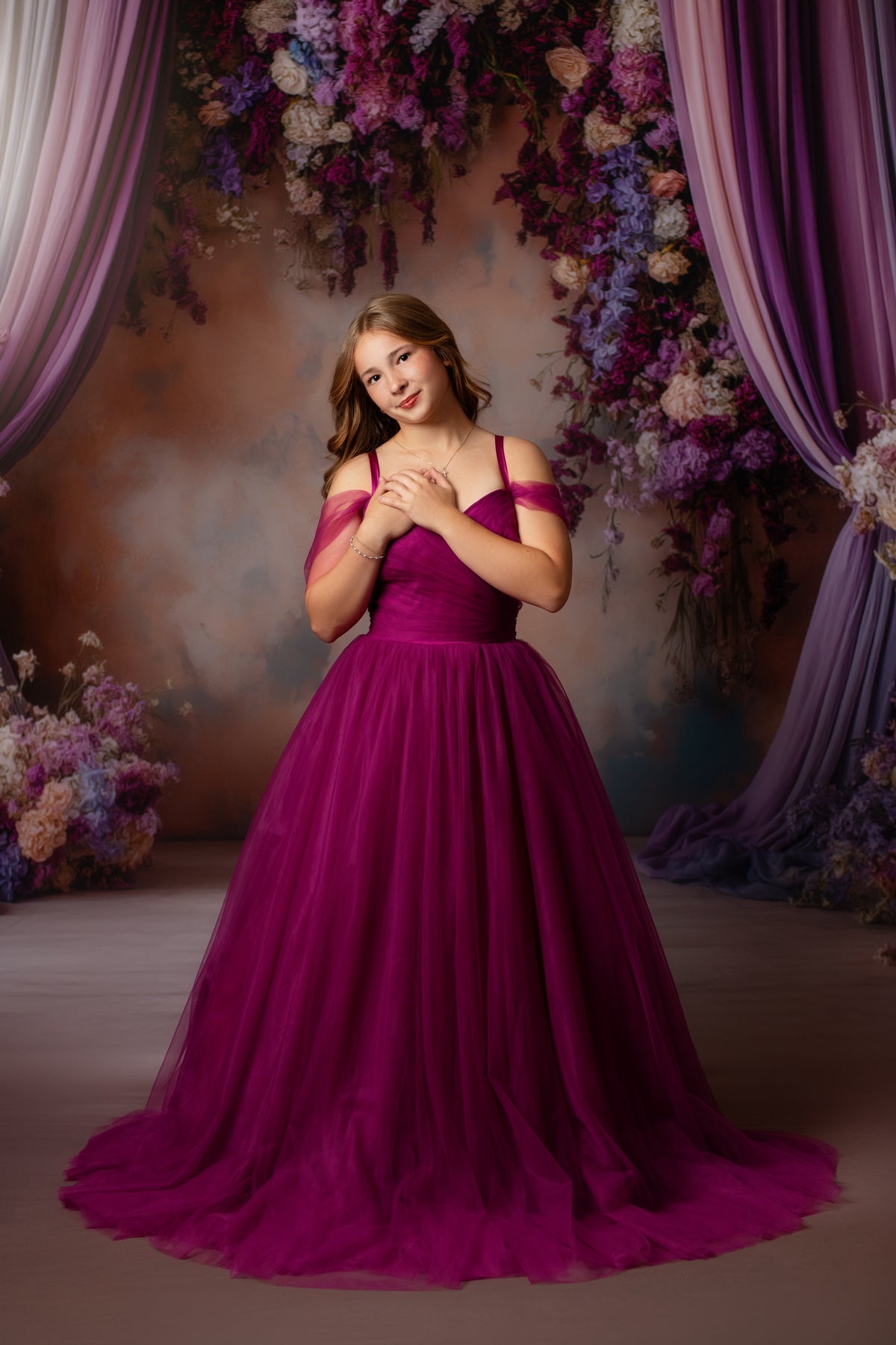 A young woman wearing a voluminous magenta ball gown with off-the-shoulder straps, standing in front of a decorative backdrop with purple drapes and floral arrangements.