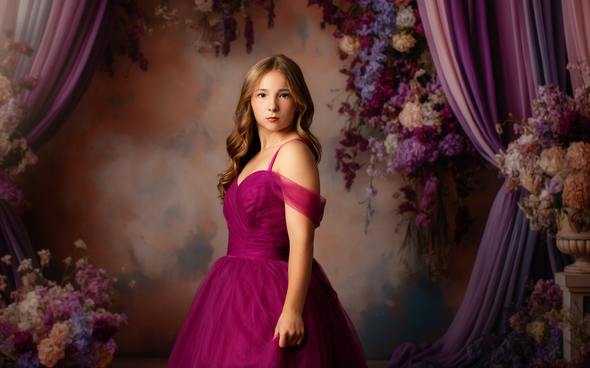 A young woman posing in a vibrant magenta off-the-shoulder ball gown against a decorative backdrop with purple drapes and floral arrangements.