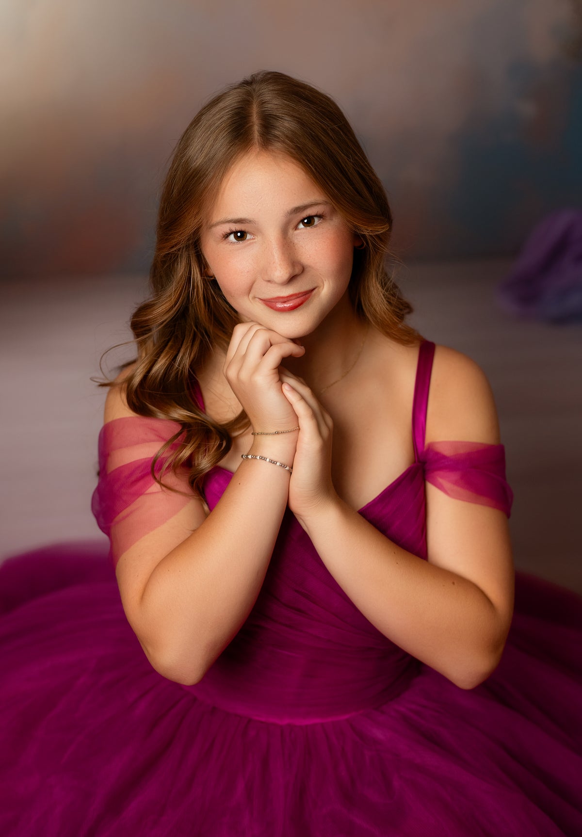 A young girl with long light brown hair wearing a magenta off-the-shoulder ball gown, posing with her hands clasped together against a soft, painterly studio backdrop.