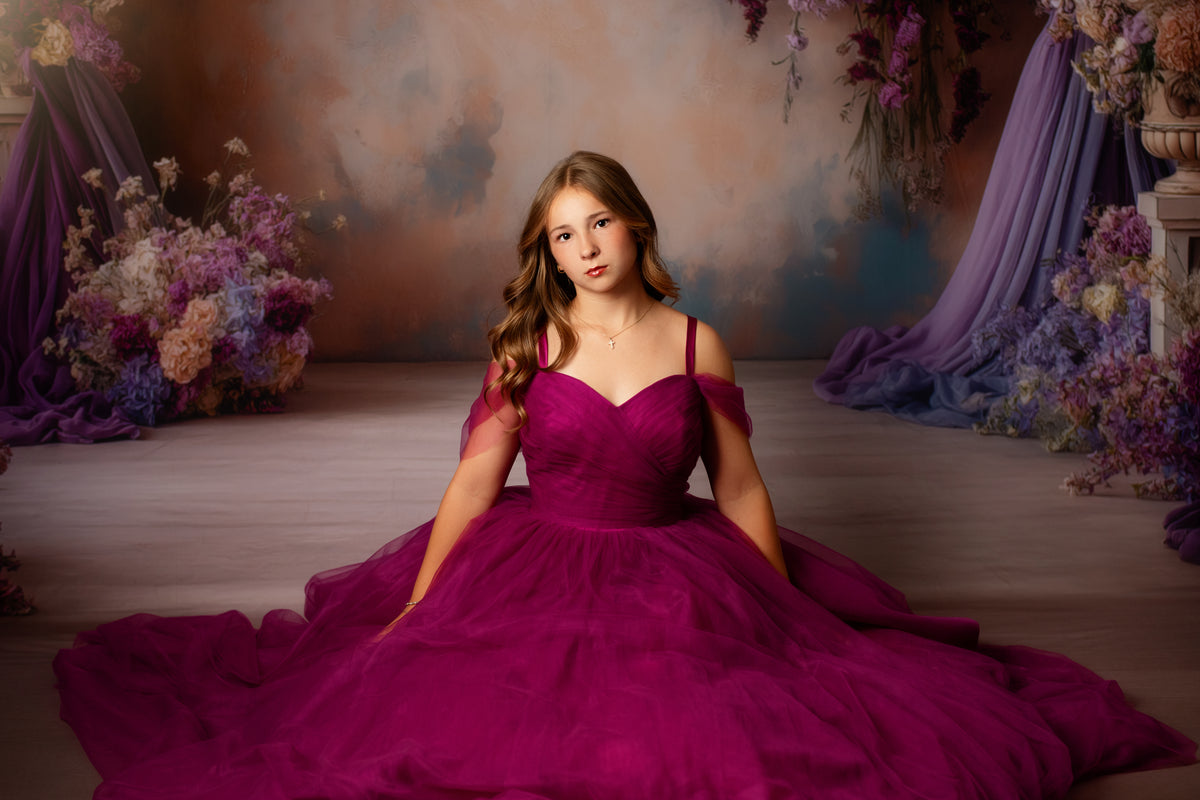 A young woman wearing a voluminous magenta ball gown sits on a wooden floor in a studio decorated with purple drapery and floral arrangements.
