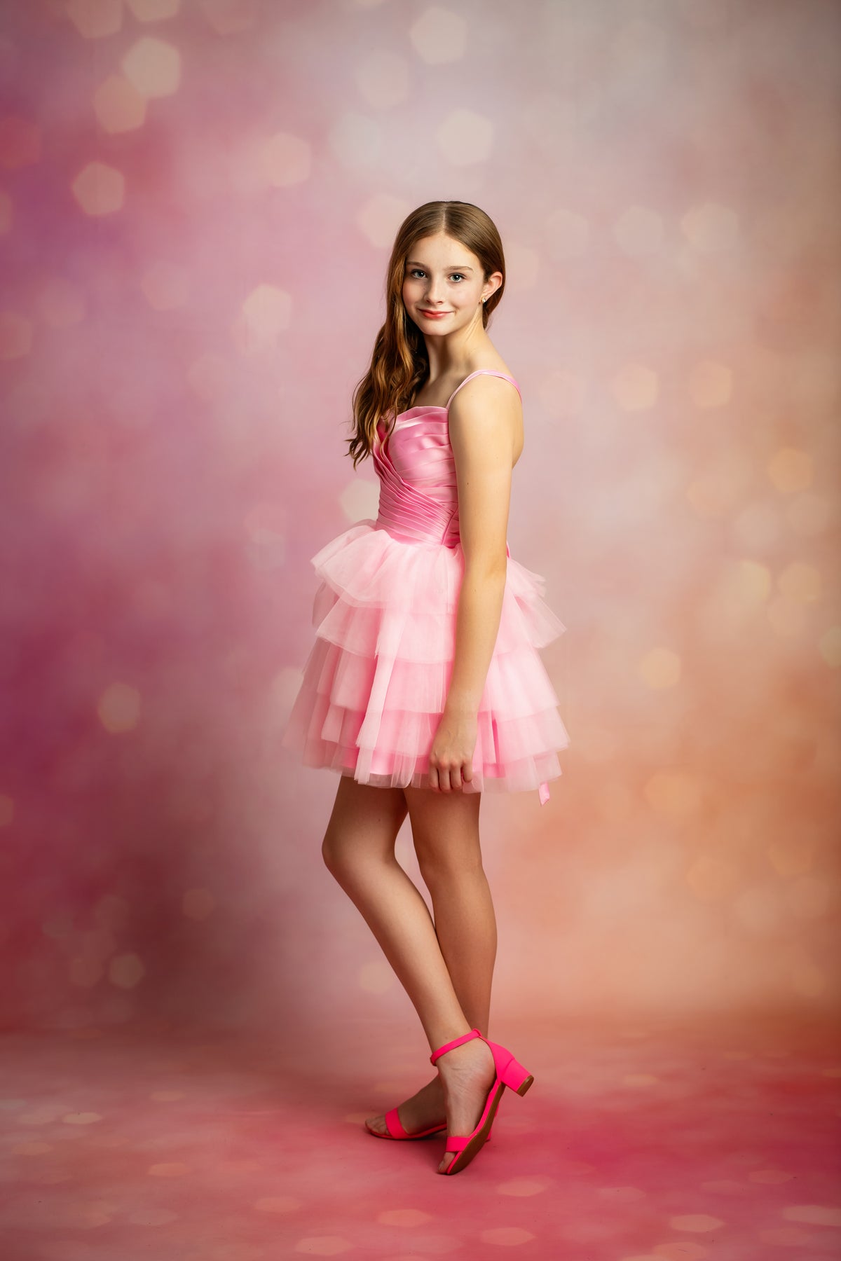 A young girl with long brown hair stands in a studio wearing a short pink tutu dress and bright pink high-heeled sandals against a pink and peach bokeh background.