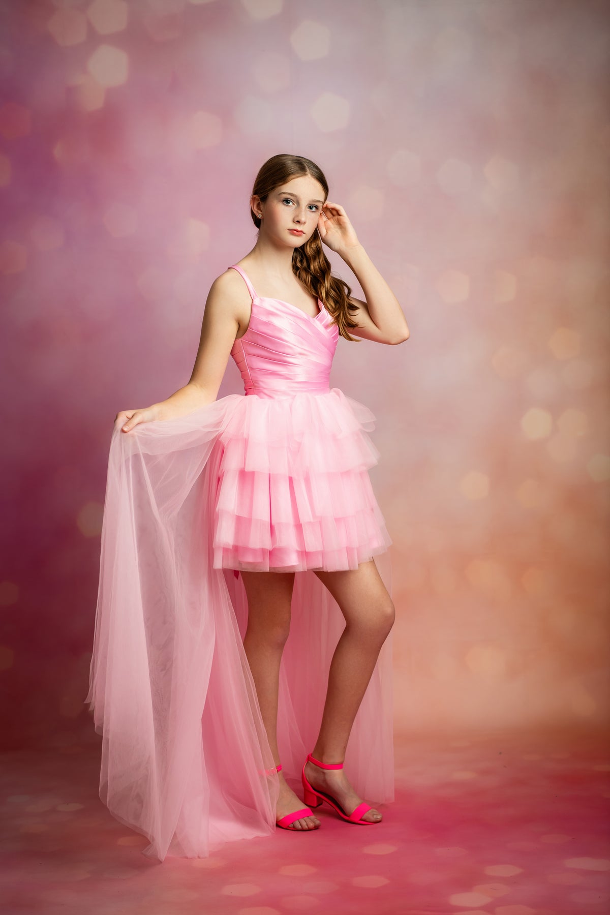 A young girl models a pink tiered tulle dress with a high-low back and sheer overskirt, paired with pink heels, against a pink bokeh studio background.