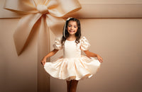 Young girl in a white dress standing next to a large beige bow on a beige background