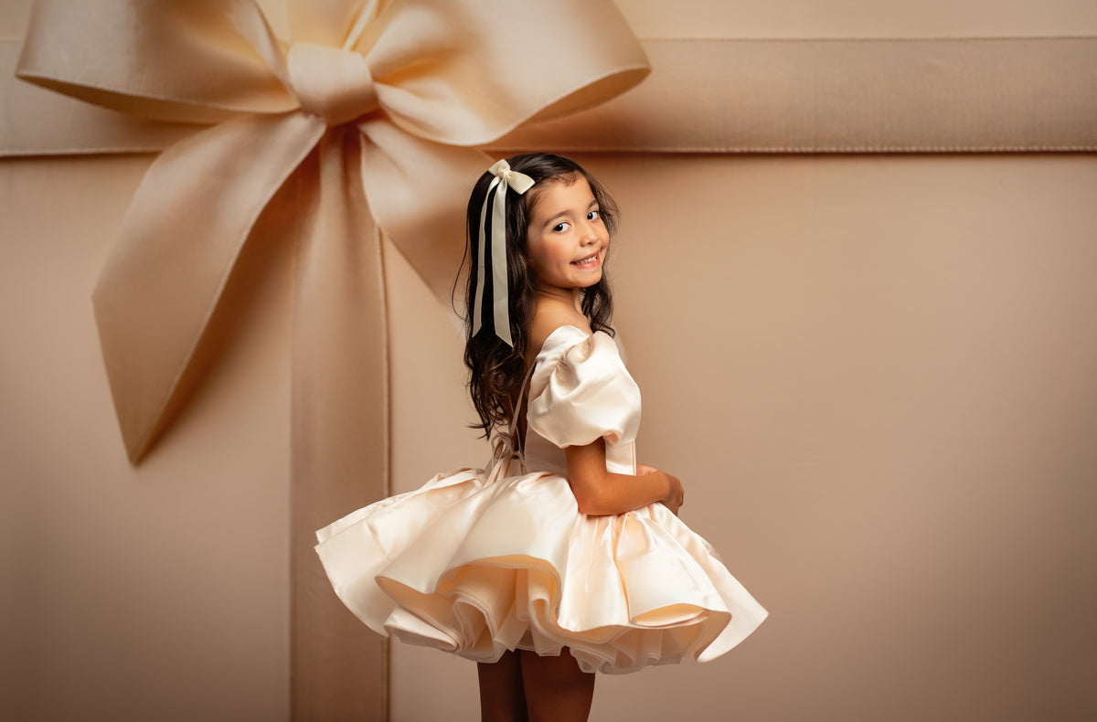 Young girl in a white dress standing next to a large gift box with a bow on a beige background