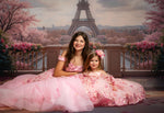 Two girls in pink dresses posing in front of a scenic backdrop with cherry blossoms and the Eiffel Tower.