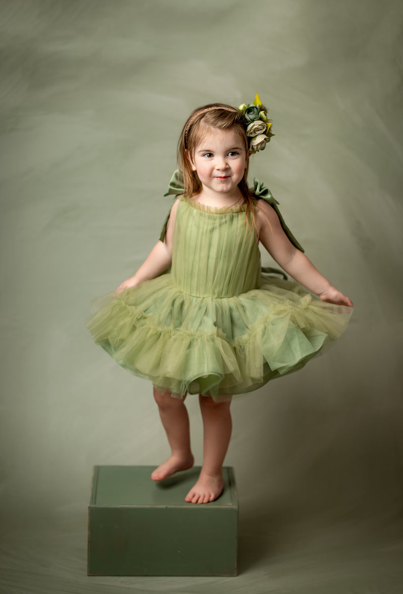 Child wearing a fairy-like olive green dress with fluttery tulle layers and statement bows, posing confidently in a photo studio setting.

