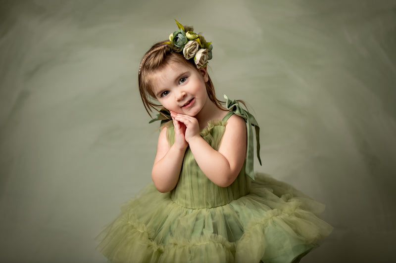 Whimsical children’s couture moment featuring a soft green ruffled dress with oversized shoulder ties and a matching floral headband.

